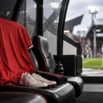 Editorial illustration of an empty red football shirt and boots on a dugout seat beside a pitch, representing a Manchester United attacking absence at Fulham.
