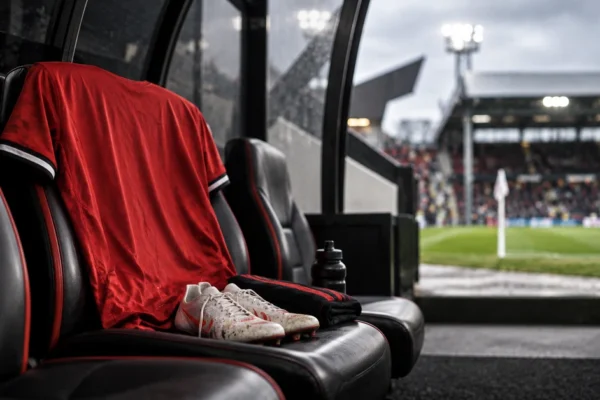 Editorial illustration of an empty red football shirt and boots on a dugout seat beside a pitch, representing a Manchester United attacking absence at Fulham.